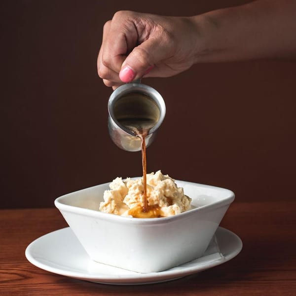 A hand pours espresso from a metal pitcher over a bowl of vanilla ice cream, an affogato dessert.