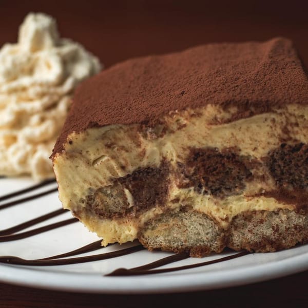 A slice of tiramisu with cocoa powder and whipped cream on a white plate with chocolate drizzle.