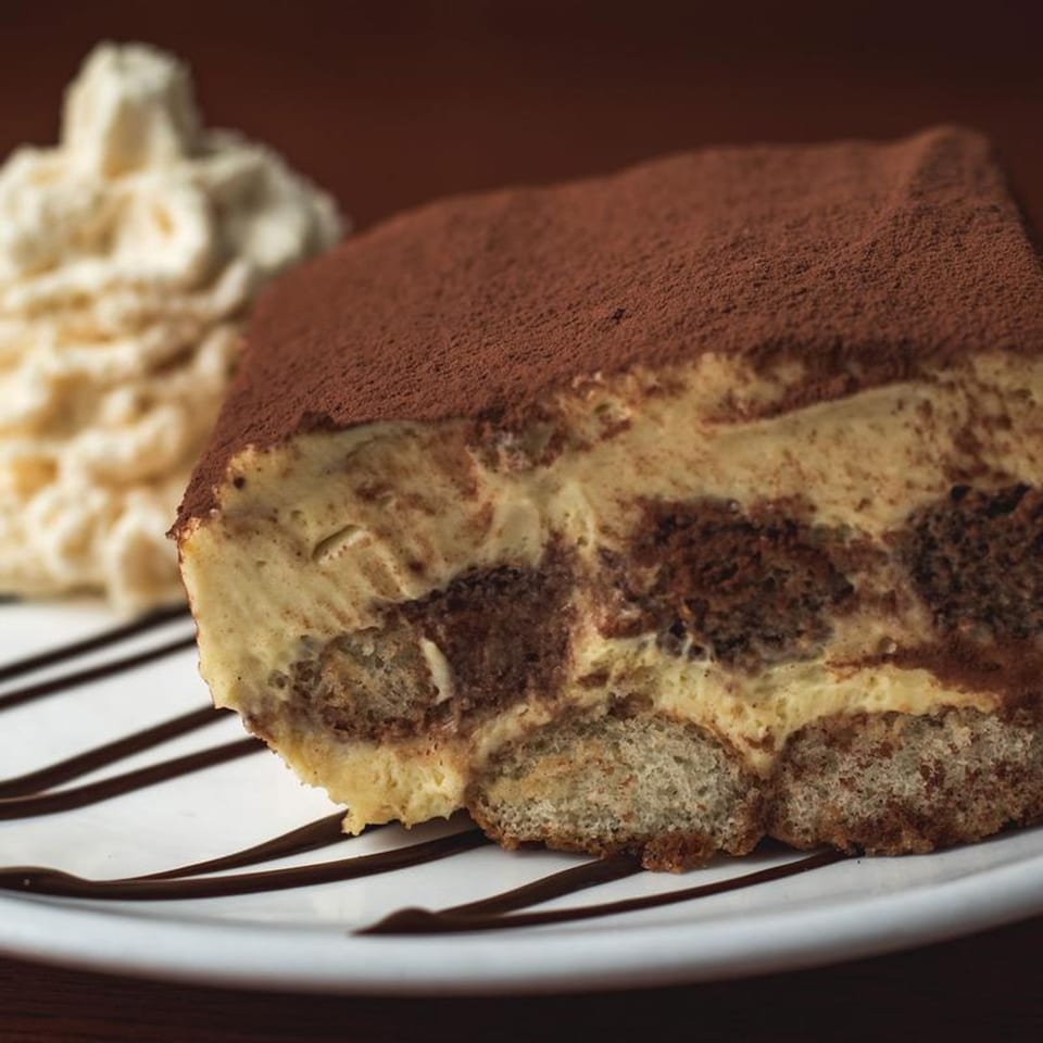 A slice of tiramisu with cocoa powder and whipped cream on a white plate with chocolate drizzle.