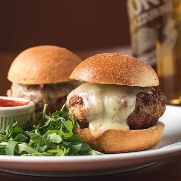 Two meatball sliders with melted cheese, served with a side of arugula and marinara sauce.